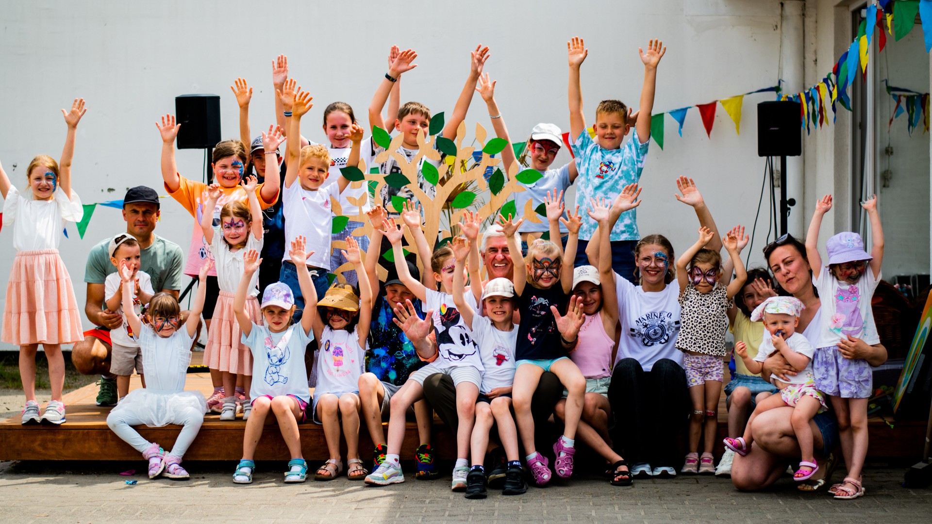 Group of children on a stage with raised hands at an outdoor festival, colorful bunting overhead and a decorative tree centerpiece.